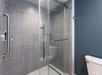 Modern bathroom with a glass shower enclosure, gray tile walls, and a white toilet. The shower features a metal handle and built-in shelf. Walls are painted a dark, contrasting color.