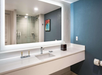 Modern bathroom with a large illuminated mirror, sink with toiletries, and a glass-enclosed shower. Blue accent wall with artwork above the toilet paper holder.