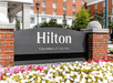 Close up of the gray and red brick sign for the Hilton Columbus at Easton with multicolored flowers in front of it on a sunny day.