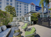 Outdoor seating area with green cushions in front of a multi-story hotel building. Tables, chairs, and umbrellas are set on the patio. Trees and plants provide greenery.