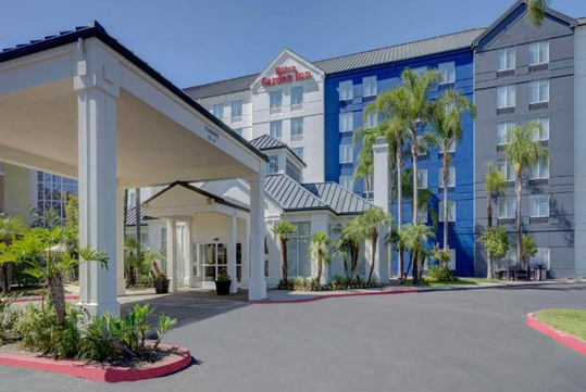 A Hilton Garden Inn hotel with a blue and white facade, palm trees, and a covered entrance, set against a clear blue sky.
