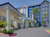 A Hilton Garden Inn hotel with a blue and white facade, palm trees, and a covered entrance, set against a clear blue sky.