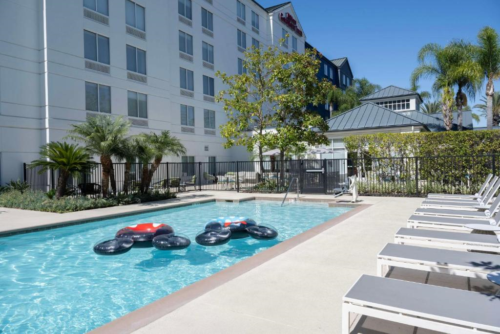 Outdoor hotel pool area with lounge chairs, inflatable float in the water, and surrounding greenery under a clear blue sky.