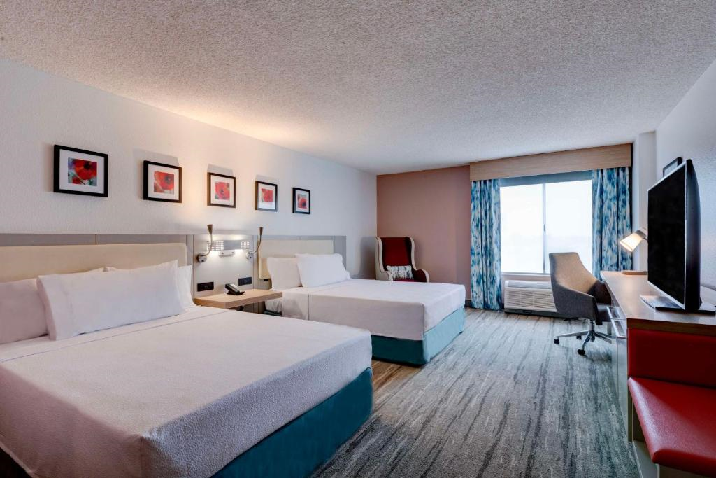 Hotel room with two beds, a TV, a desk with a chair, and a large window with curtains. Walls feature framed artwork. Room is decorated in neutral tones with pops of color.