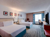 Hotel room with two beds, a TV, a desk with a chair, and a large window with curtains. Walls feature framed artwork. Room is decorated in neutral tones with pops of color.