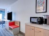 Hotel room with a TV on a wooden cabinet, a red bench, a microwave, and a coffee maker on a countertop. A colorful painting is on the wall above.