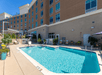 Outdoor hotel pool area with lounge chairs, umbrellas, and plants. A multi-story brick building is in the background under a clear blue sky.