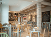 Café interior with wooden tables and chairs. Walls feature murals and chalkboard menus. Ceiling lights are hanging, creating a warm atmosphere.
