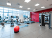A gym with treadmills, ellipticals, a weight machine, and a red exercise ball on a tiled floor. Large windows provide natural light.