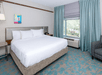Hotel room with a large bed, white bedding, and teal accents. A window with a view, curtains, a chair, bedside tables, a small painting, and a wall lamp are visible.