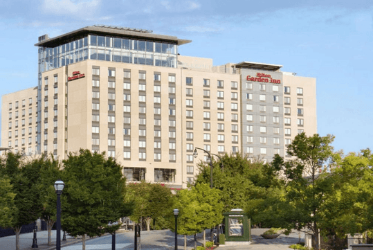 Exterior view of a multi-story Hilton Garden Inn hotel, featuring large windows and a light-colored facade, with trees and lamp posts in the foreground.