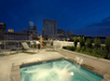 Rooftop pool with water features overlooking a city skyline at night, surrounded by lounge chairs and green plants.