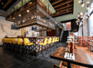 Restaurant interior with a central bar, yellow barstools, and tiled design. Wooden tables and chairs are set nearby. A staircase leads to the upper level, and a wall map decorates the space.