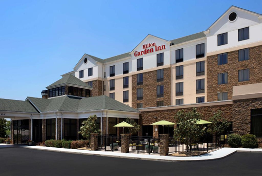 Exterior view of a Hilton Garden Inn with a stone facade, multiple windows, and a small outdoor patio with green umbrellas.