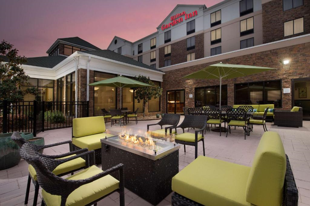 Hotel courtyard with patio furniture around a lit fire pit, green cushions, and umbrellas. The hotel's exterior and signage are visible in the background under a twilight sky.