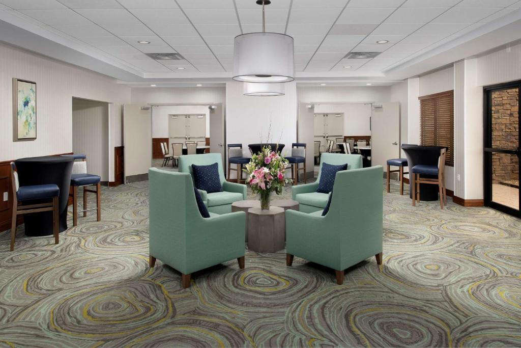 A waiting area with patterned carpet, teal armchairs, a round table with flowers, and a large ceiling light. Tables and chairs line the walls in the background.