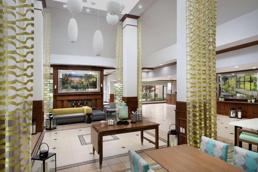 Spacious hotel lobby with modern decor, featuring patterned columns, contemporary seating, artwork, and large light fixtures. A central wooden table with refreshments is visible.