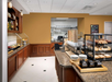 Hotel breakfast area with cereal dispensers, pastries, bread, and chafing dishes on a counter. Shelving with condiments and tableware in the background. Kitchen visible through an open doorway.