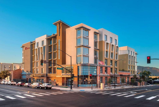 Street view of a modern multi-story hotel at an intersection with clear skies. An American flag is displayed, and cars are parked along the street.