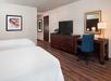 Hotel room with two beds, a wooden dresser with a TV, a desk with a blue chair, a lamp, and wall art of horses. The room has hardwood flooring and neutral-colored walls.