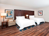 A hotel room with two double beds, a beige armchair, two framed artworks, and wooden flooring. There are two lamps on the bedside tables.
