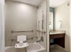 A single lighted vanity mirror, fresh towels, bath chair, shower and tub with grab bars in an accessible guest bathroom.