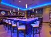 A "U" shaped bar with white counter tops and chairs and purple lights shining down on them plus a wall of liquor.