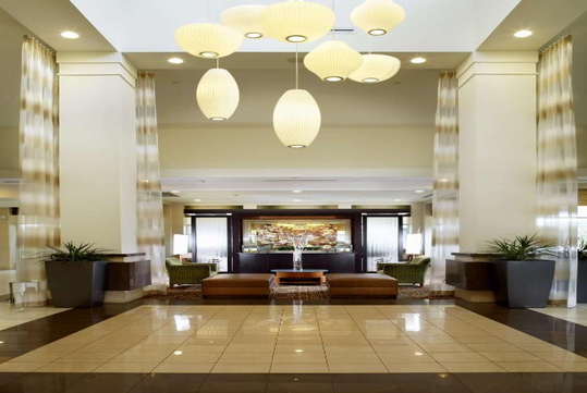 Hotel lobby with hanging oval lights, beige and brown tones, tall plants, and a seating area in front of a colorful wall art piece.