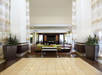 Spacious hotel lobby with tall ceilings, large potted plants, a tile floor, and a seating area with a TV in the background.