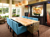 A dining room with a large wooden table, surrounded by turquoise and patterned chairs. Pendant lights hang above, and a decorative display is in the background.