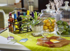 A table with bottles of wine in a rack, a potted plant, glass jars of yellow and green fruits, two empty wine glasses, and four colorful tags on a green tablecloth.