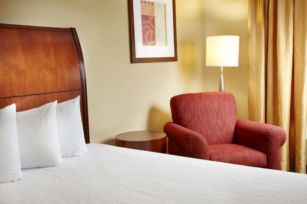 Hotel room with a large bed, three pillows, a red armchair, a wooden side table, a tall lamp, and artwork on a cream-colored wall.