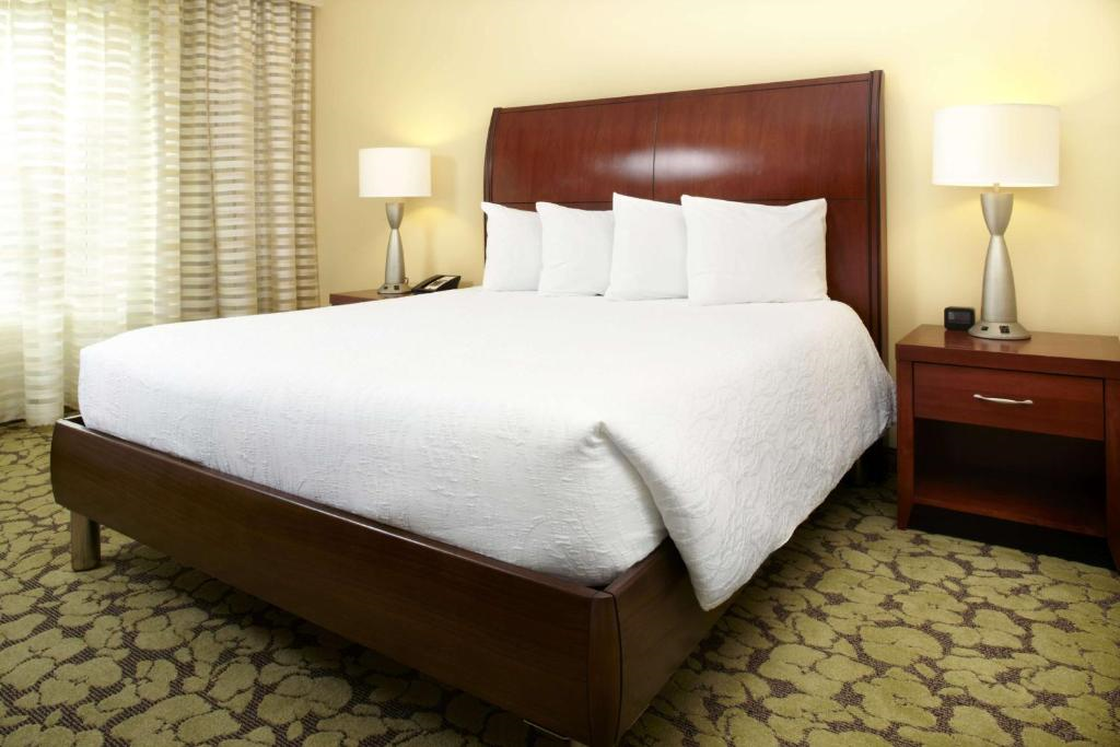 A neatly made bed with four white pillows, dark wooden headboard, and two bedside tables with lamps. The room has carpeted flooring and a window with patterned curtains.