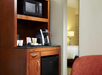 Hotel room kitchenette with a microwave, coffee maker, and mini fridge on a wooden cabinet. Part of a bed and a lamp are visible in the adjacent room.