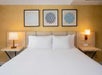 Neatly made bed with white linens, flanked by two lamps on wooden nightstands. Three framed geometric art pieces hang on a yellow wall above the headboard.