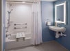 Accessible bathroom with a shower-tub combo featuring grab bars and a shower chair. A sink with a backlit mirror is mounted on a blue wall. A white curtain separates the shower area.