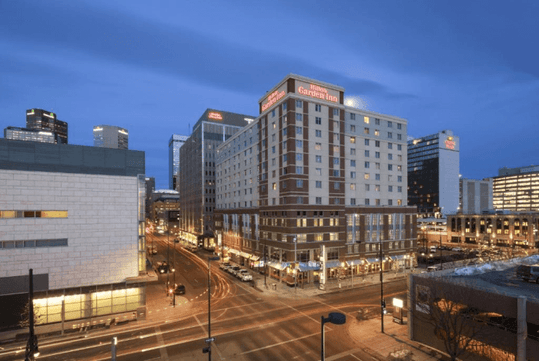 Wide shot of the exterior of the Hilton Garden Inn Denver Downtown on a cloudy night with cars zooming by.