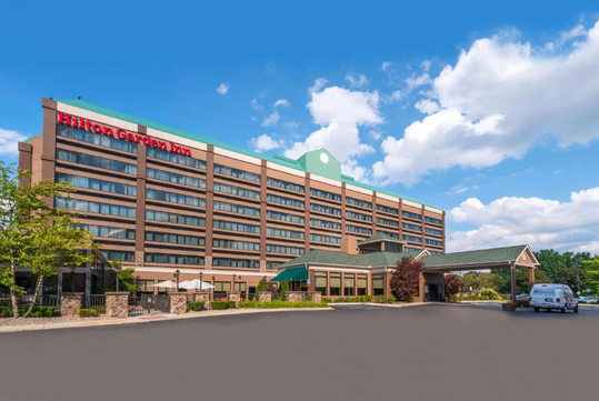 A large hotel with multiple floors, featuring a sign that reads "Hilton Garden Inn." The building is surrounded by a parking lot and greenery, under a partly cloudy sky.