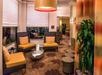 Hotel lobby with wicker chairs and orange cushions, a metallic coffee table, potted plant, and floral carpet. Large windows with white blinds and a hanging light fixture are visible.