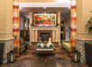 Hotel lobby with a fireplace, seating area featuring patterned chairs and a green sofa, a central table with plants, pendant lamps, and vibrant artwork above the mantel.