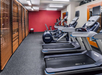 A gym room with treadmills and elliptical machines on the right. Brown blinds cover the windows on the left, and a red accent wall is visible at the back.