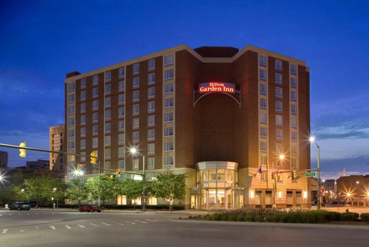 A multi-story Hilton Garden Inn hotel at dusk, with illuminated signs and building lights, on a street corner surrounded by trees and streetlamps.