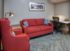 A room with a red sofa, matching armchair, and a side table. A framed picture hangs above. A desk with a mesh chair and lamp is in the background.