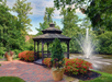 A wooden gazebo stands beside a pond with a spraying fountain. Red flowers and green foliage surround the brick pathway leading to the gazebo.