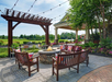 Outdoor patio with wooden benches surrounding a fire pit, string lights above, and colorful cushions. A pergola and landscaped garden are in the background.