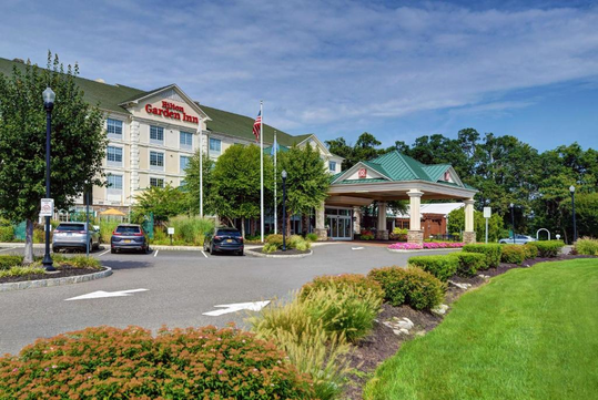 Front view of a Hilton Garden Inn hotel with a green roof, a driveway, parked cars, and landscaped greenery under a blue sky.