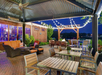 Outdoor patio with wooden tables and chairs, a pergola with string lights, and a fire pit. Lounge seating is visible near a wooden building with large windows.