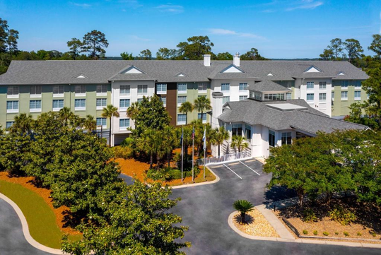Aerial view of a three-story hotel surrounded by trees and a parking lot, with multiple windows and a grey roof. Several flags are displayed near the entrance.