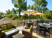 Outdoor patio with wicker furniture, green cushions, round tables, and umbrellas. Surrounded by trees, sunny weather.