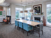Dining area with a wooden table, eight chairs, three pendant lights, and a fireplace. Large windows and art decorate the room, providing a bright, modern atmosphere.